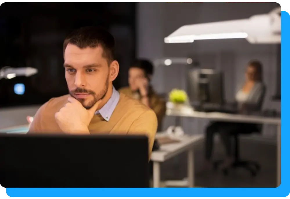 A man sits at a desk working on a laptop, in a dim office with colleagues blurred in the background.